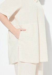 Parte superior color crema con finas líneas de color marrón claro; mangas cortas y un corte relajado. Cuenta con un bolsillo lateral y una textura suave y lisa.