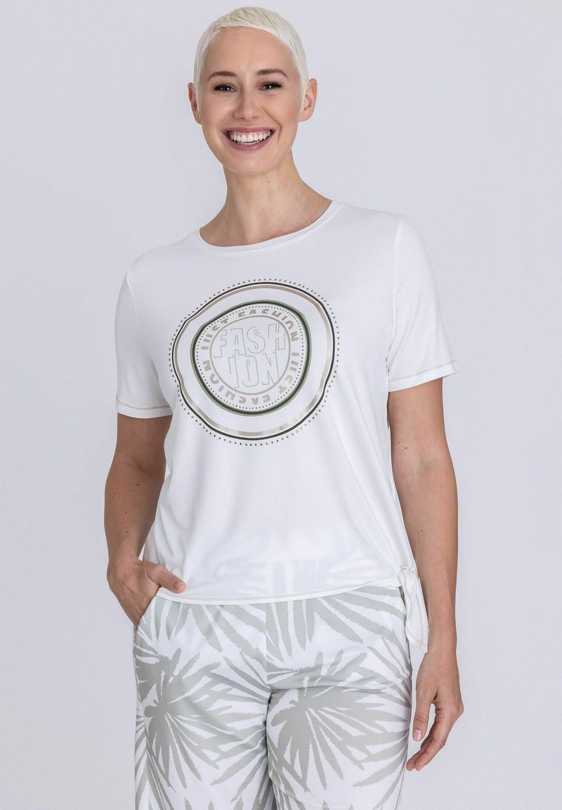 Vrouw met kort blond haar, gekleed in een wit T-shirt met opdruk en witte broek met grijze palmbladpatronen, glimlachend met één hand in haar zak.