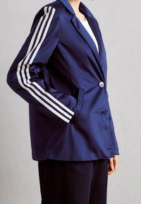 Tmavě modré sako má jeden knoflík, dvě boční kapsy a bílé třípruhové ozdoby na rukávech. Hladká látka s přizpůsobeným střihem.