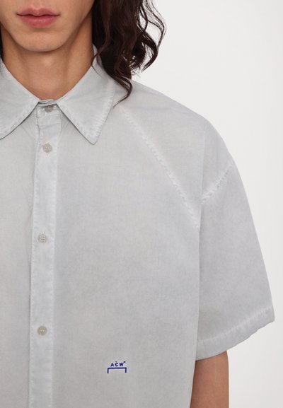 Chemise à manches courtes gris clair avec un col boutonné, un léger éclat et un logo brodé bleu sur la poitrine. Texture lisse et coupe ajustée.