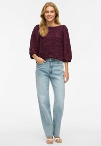 Žena v texturované tmavě fialové blůze s nabíranými rukávy, světle modrých kalhotách s vysokým pasem a béžových sandálech, stojící s jednou rukou v kapse.