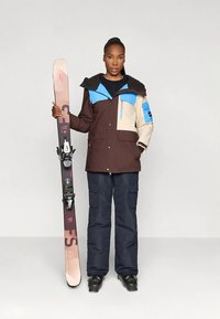 Vrouw in een kleurblok ski-jack en -broek, die een roze en zwarte snowboard vasthoudt, staand tegen een effen lichtgrijze achtergrond.