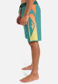 Bañador en color verde azulado con acentos en gradiente naranja, con una cintura elástica ajustable y diseño de logo en el costado. Fabricado con material liviano y de secado rápido.