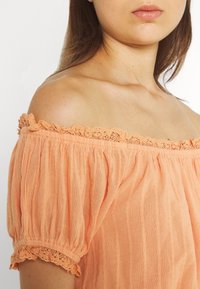 Blusa pescoço a spalle scoperte in pesco, con tessuto testurizzato, bordi in pizzo arricciati sulle maniche e sottili righe verticali.
