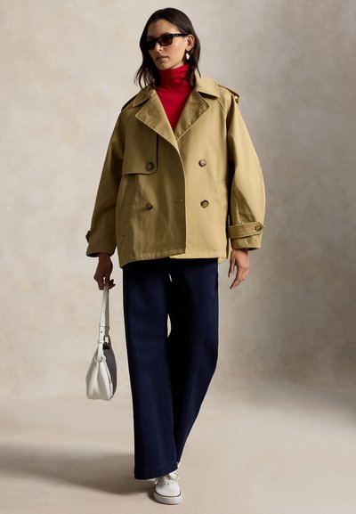 Γυναίκα με υπερμεγέθη δίπλα κουμπωμένο μπεζ σακάκι, κόκκινο ζιβάγκο, ναυτικό μπλε παντελόνι φαρδιάς γραμμής, λευκά αθλητικά παπούτσια, μαύρα γυαλιά ηλίου, κρατώντας λευκή τσάντα.