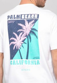 T-shirt en coton blanc avec un imprimé graphique de palmiers roses sur fond bleu avec le texte "Palm Beach" et "Californie, Los Angeles."