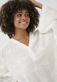 Blusa bianca con ricami floreali, scollatura a V e dettagli con bottoni. Il tessuto ha un aspetto morbido e texturato con maniche ampie.