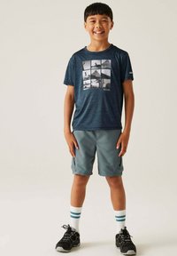 Marineblaues T-Shirt mit kurzen Ärmeln, das ein Gittermuster aus schwarz-weißen Outdoor-Bildern zeigt; kombiniert mit grauen Shorts und schwarzen Sportschuhen.