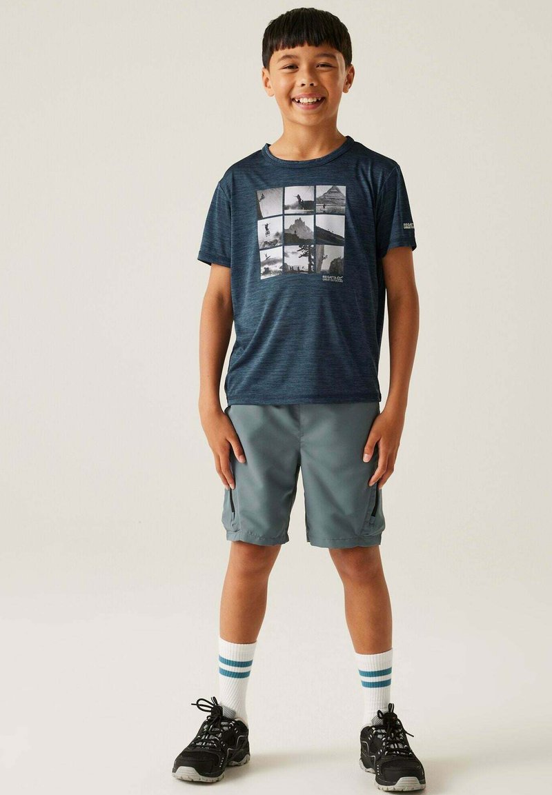 Marineblaues T-Shirt mit kurzen Ärmeln, das ein Gittermuster aus schwarz-weißen Outdoor-Bildern zeigt; kombiniert mit grauen Shorts und schwarzen Sportschuhen.