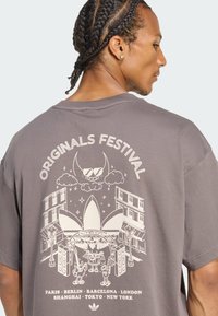 Gray oversized t-shirt featuring a graphic print on the back with "Originals Festival" text and city names, smooth cotton texture.