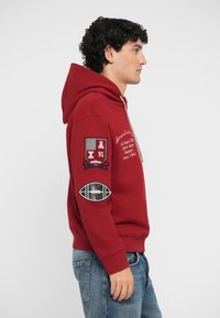 Rød hoodie med snor og lomme foran. Har broderede lapper på ærmerne, herunder sportsrelaterede designs. Materiale i bomuldsblanding.