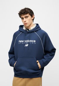 Jonge man draagt een marineblauwe New Balance Athletics hoodie met handen in de buidelzak, gecombineerd met beige broek, staand tegen een effen achtergrond.