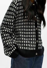 Melnā un baltā rakstā pārklāta jaka ar pogu aizdari, ribotu apakšmalu un mīkstu tekstūru; ar izteiktu kvadrātveida dizainu un apaļām metāla pogām.