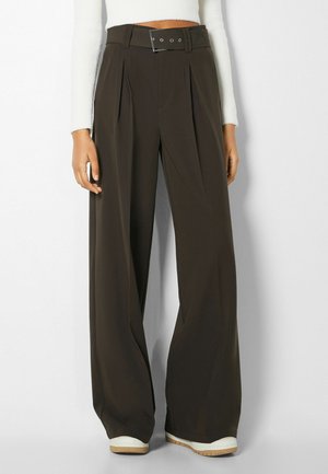 Kobieta ubrana w ciemnobrązowe spodnie z szerokimi nogawkami i wysokim stanem, ze skórzanym paskiem w tym samym kolorze, oraz białe trampki, w połączeniu z białym, krótkim topem z długim rękawem.