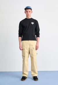 Svart langermet skjorte med hjerte-logo, beige cargo bukser med lommer, og svarte sko med gummisåler. Blå caps fullfører antrekket.