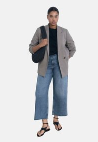 Blejzer s uzorkom houndstooth, s jednim gumbom, uparen s širokim svijetloplavim trapericama i crnim ravnim sandalama, drži teksturiranu crnu torbicu.