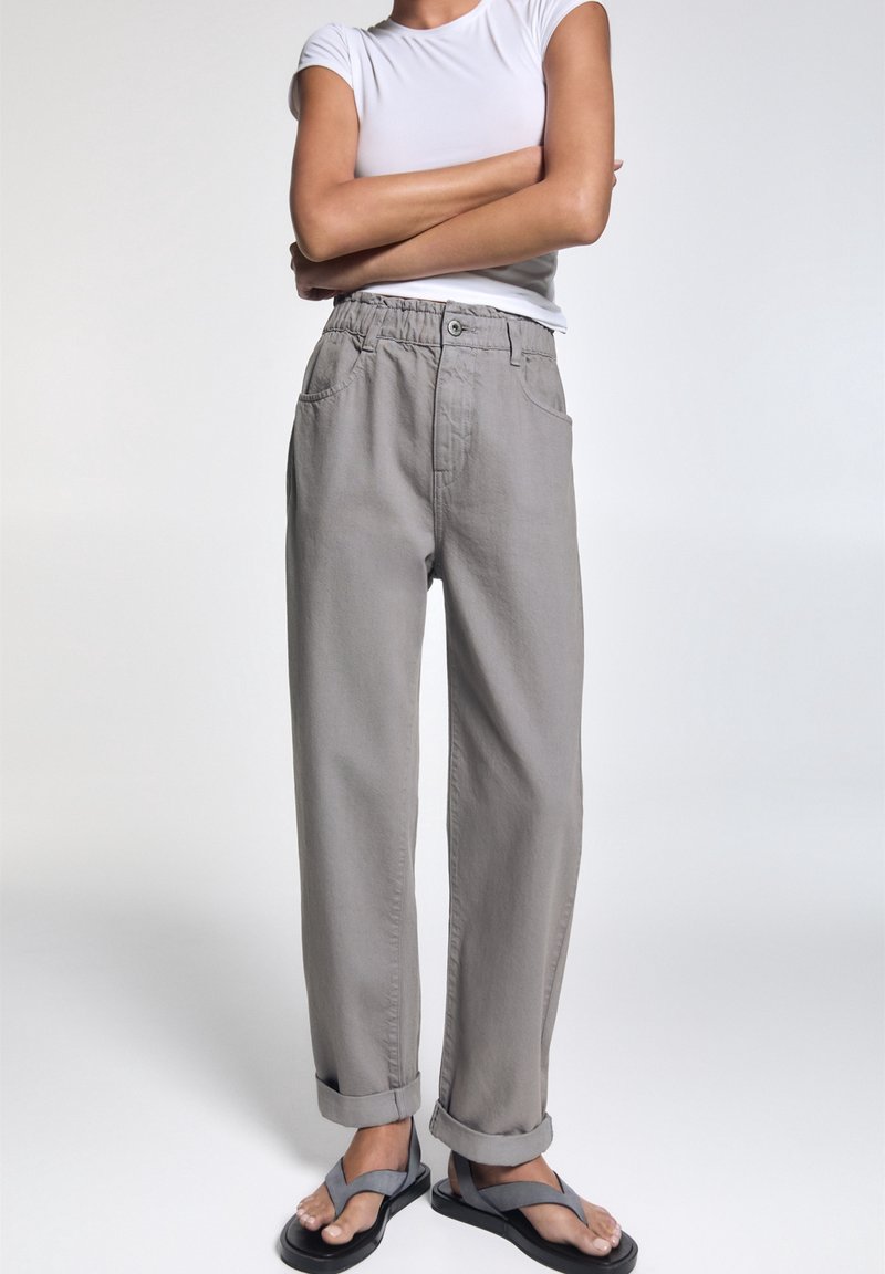 Personne debout, bras croisés, portant une chemise blanche à manches courtes, un pantalon gris ample avec les revers remontés et des sandales grises.