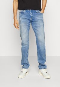 Jeans de lavado claro en azul con un corte recto, que presentan un ligero desvanecimiento, cinco bolsillos y cierre de cremallera. Llevados con zapatillas blancas.