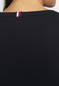 Gros plan du haut du dos d'une personne portant un haut noir côtelé avec une étiquette en tissu rouge, blanc et bleu au niveau de l'encolure.