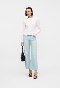 Γυναίκα με ανοιχτό ροζ ζακέτα με κουμπιά, φαρδιά ανοιχτό μπλε τζιν, μαύρες γόβες, κρατώντας μαύρη τσάντα χειρός με το ένα χέρι στην τσέπη.