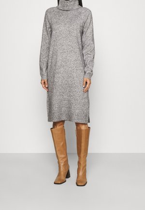 Femme portant une robe en maille gris à manches longues et col roulé, associée à des bottes en cuir marron clair montant jusqu'aux genoux, debout devant un fond uni.