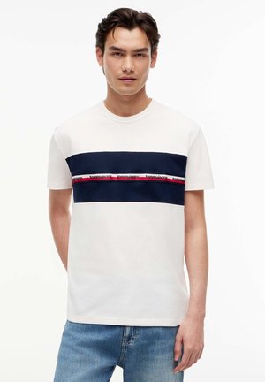 Junger Mann, der ein weißes T-Shirt mit einem dunkelblauen und roten horizontalen Streifen über der Brust und hellblaue Jeans trägt, steht vor weißem Hintergrund.