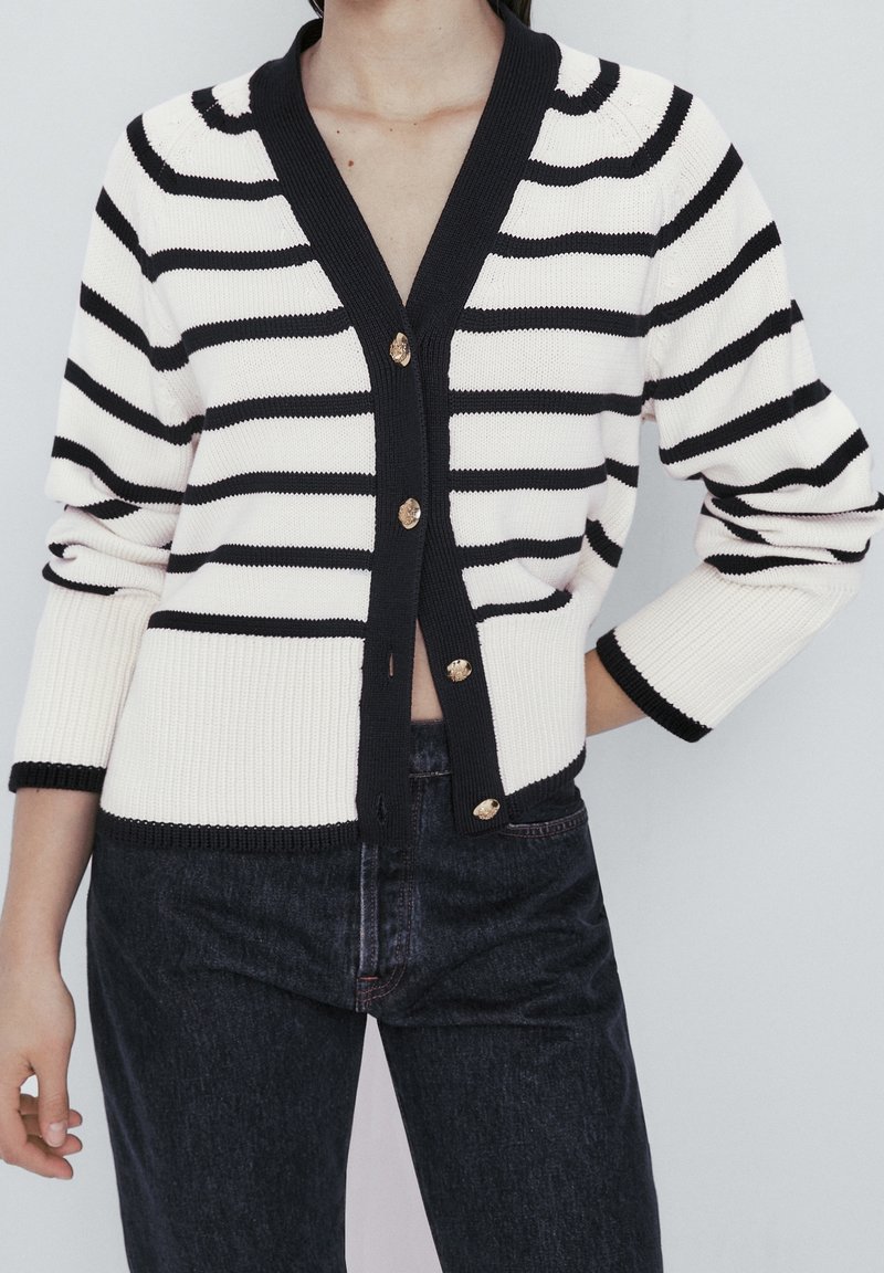Femme portant un cardigan rayé noir et blanc boutonné avec des bordures noires et un jean foncé taille haute sur un fond uni.