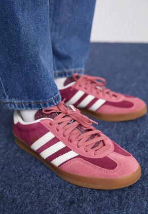 Zapatillas de ante rosa con rayas blancas y diseño de cordones, que cuentan con una puntera texturizada y suela de goma. Llevadas con vaqueros azules.