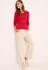 Rotes Baumwoll-Langarmshirt mit V-Ausschnitt, beige weite Hose mit Taschen und hellen Sneakers, vor einem weißen Hintergrund stehend.