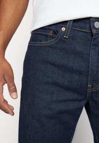 Tmavě modré džíny ze džínoviny s úzkým střihem, zdobené měděnými kovovými prvky a žlutým prošíváním. Detail pásu a ruky položené na něm.