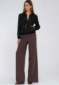 Czarna kurtka bomberka z cekinami, zapinana na zamek, z prążkowanymi mankietami. Połączona z szerokimi brązowymi spodniami i butami na obcasie. Proste tło.