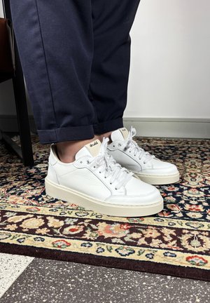 Witte leren sneakers met beige zolen gedragen met donkerblauwe omgeslagen broekspijpen, staand op een bontgekleurd patroon vloerkleed naast een zwarte stoel.
