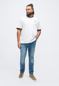 Homme aux dreadlocks portant un t-shirt blanc avec une bordure noire, un jean bleu clair et des baskets beiges, debout avec une main dans la poche devant un fond blanc.