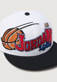 Bijela bejzbol kapa s crnim rubom, sa izvezenim "Jordan" natpisom u crvenoj i plavoj boji, grafikonom košarkaške lopte i detaljem "1984".