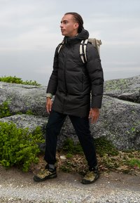 Polstret sort jakke med høj krave, parret med mørke bukser og grønne vandresko. En lys rygsæk bæres over skuldrene.