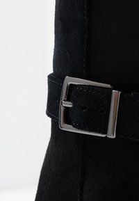 Zoom su un cinturino di stivale in suede nero con fibbia rettangolare argentata su sfondo bianco.