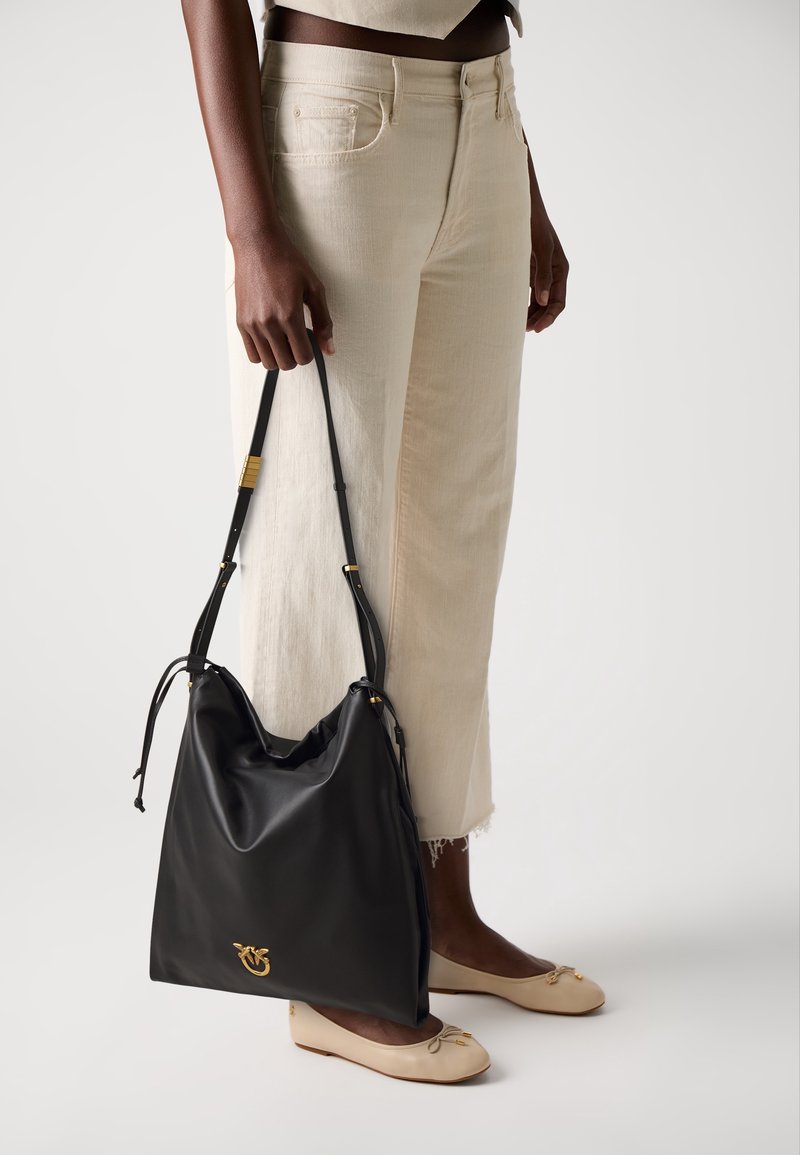 Personne portant un pantalon beige clair coupé et des ballerines crème tenant un grand sac à main en cuir noir avec des détails dorés du logo.