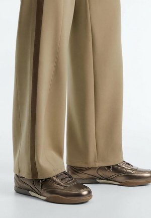 Egy személy lábainak hosszirányú képe, aki bézs színű bő szárú nadrágot visel barna oldalsávokkal és fémes bronz színű tornacipőket világos padlón.