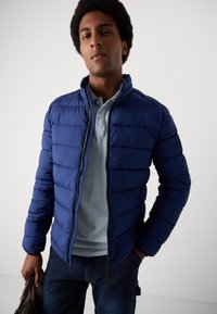 Marinefarbener Puffer-Jacke mit horizontalen Nähten, hohem Kragen und Reißverschluss. Kombiniert mit einem hellblauen Polo darunter und Jeans.