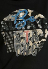 Schwarzes T-Shirt mit einem runden, Tierdruckdesign. Mit dem Schriftzug "Be STRONG" in metallischen, genähten Buchstaben, akzentuiert mit blauen und roten Elementen.
