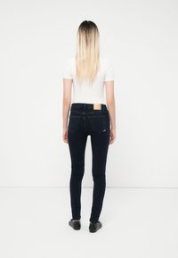 Sieviete ar gariem blondiem matiem stāv vērsta pret baltu sienu, ģērbusies baltā t-kreklā, tumšās cieši pieguļošās džinsās un melnās kurpēs.