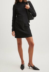 Donna che indossa un mini abito aderente nero a maniche lunghe, décolleté neri con tacco kitten e porta una borsa a spalla nera.