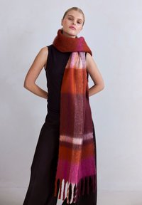 Wollen sjaal in levendige tinten van rood, roze en paars met een ruitpatroon en franjes aan de uiteinden, om de nek gedrapeerd. Zachte textuur.