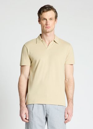Homme portant une chemise beige à manches courtes avec col et un pantalon rayé bleu clair et blanc, debout devant un fond blanc uni.