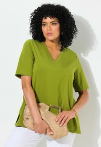 Top verde oliva a maniche corte con scollatura a V e vestibilità rilassata, con spacchetti laterali, abbinato a una pochette metallica oro. Sfondo con muro neutro.