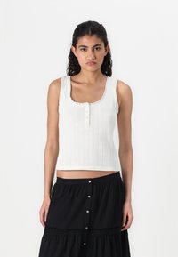 White ribbed tank top with a square neckline, decorative scalloped edges, and three buttons down the front. Styled with a black skirt.