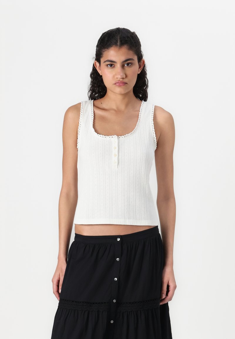 White ribbed tank top with a square neckline, decorative scalloped edges, and three buttons down the front. Styled with a black skirt.