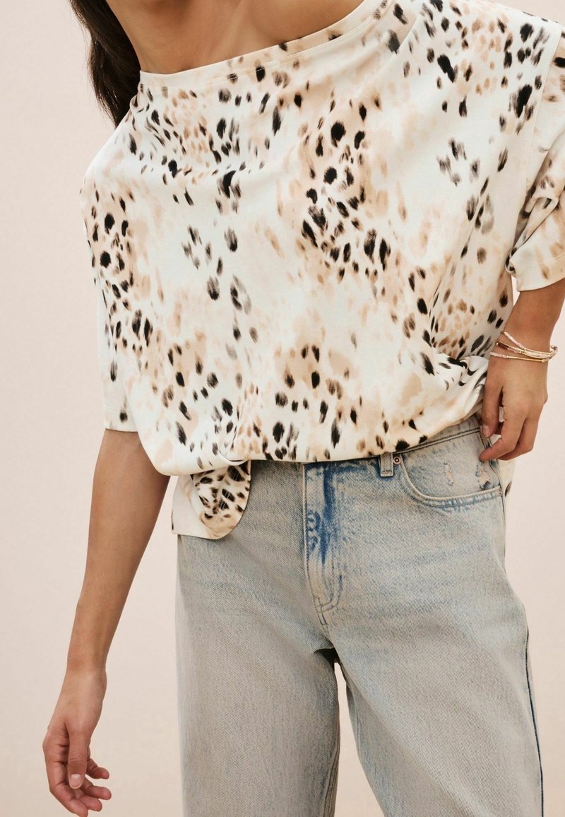 Femme portant un haut blanc ample avec des taches noires et beiges, rentré dans un jean en denim bleu clair taille haute, avec des bracelets au poignet droit.