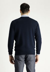 Dunkelblauer Pullover mit gerippten Bündchen und Saum, aus weichem Strickmaterial. Getragen über einem hellblauen Hemd mit Kragen, das ein minimalistisches Design aufweist.
