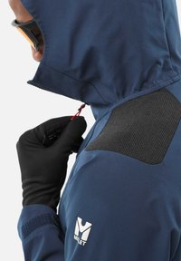 Personne portant une veste Millet bleue avec une capuche, ajustant un tirette rouge, portant des gants noirs et des lunettes noires.